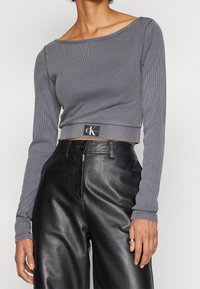 Grå ribbad långärmad crop top med båt-halsringning och en CK Jeans-logotyp vid nederkanten, kombinerad med svarta löst sittande läderbyxor.