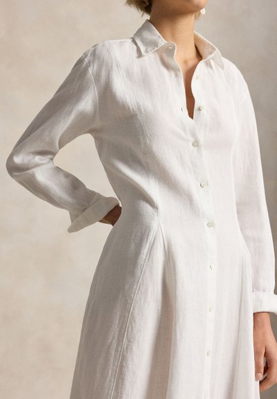 Γυναίκα που φορεί μακρυμάνικο λουλουδένιο λευκό φόρεμα με κουμπιά και γιακά, χέρι στη μέση, ουδέτερο φόντο.