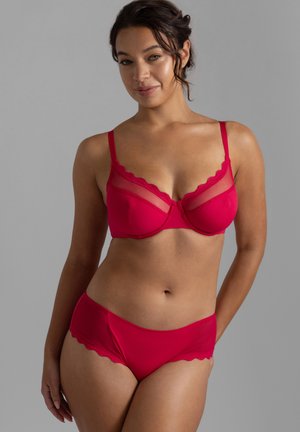 Frau mit dunklem Haar, die einen roten BH und passende Spitzenunterwäsche trägt und vor einem schlichten grauen Hintergrund steht.