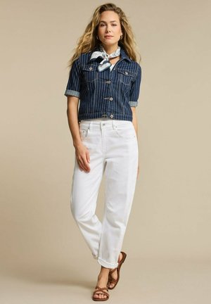 Vrouw draagt een blauw, met fijne strepen gestreepte jas, witte broek, bruine sandalen en een wit sjaaltje met patroon, staand tegen een beige achtergrond.