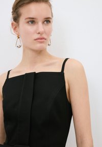 Schwarzes ärmelloses Kleid mit geradem Ausschnitt und einer zentralen Faltenzier. Das Model trägt runde, metallische Ohrringe mit einem Drahtdesign.