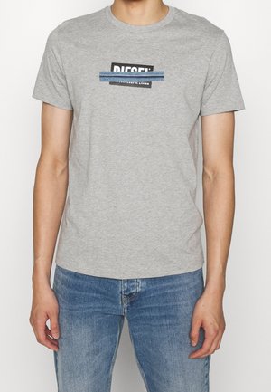 Mężczyzna ubrany w jasnoszary t-shirt z krótkim rękawem, z częściowo zasłoniętym poziomym denimowym paskiem logo Diesel w kolorach czarnym i białym, w zestawie z niebieskimi dżinsami.