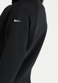 Černý sportovní top s vysokým límcem, hladkou texturou a bílým logem Nike na levém rukávu. Elegantní, přiléhavý střih.