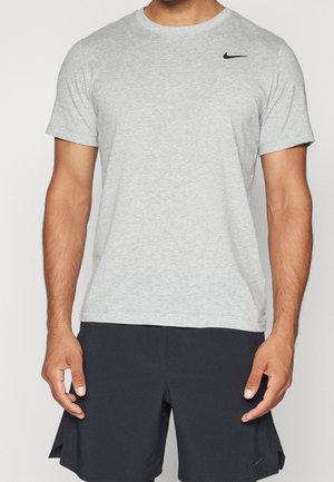 Man draagt een lichtgrijs Nike T-shirt met korte mouwen en zwarte Nike shorts, staat met ontspannen armen langs het lichaam, neutrale achtergrond.