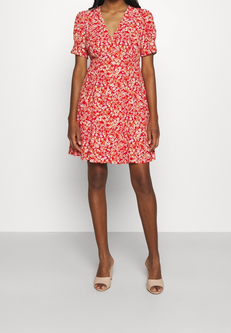 Femme portant une robe portefeuille courte rouge à fleurs avec manches courtes et des sandales beiges ouvertes à l'avant avec un talon bloc, debout sur un fond blanc.