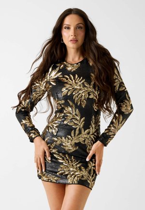 Donna con capelli lunghi e scuri che indossa un mini abito nero decorato con motivi di foglie dorate in paillettes, in posa con le mani sui fianchi su uno sfondo bianco.