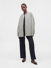 Personne portant une veste boutonnée gris clair oversize, un pantalon noir à jambes larges et des ballerines noires pointues, debout sur un fond blanc.