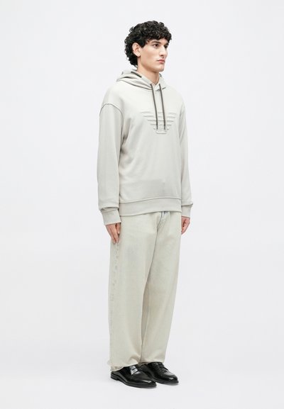 Ανοιχτό γκρι φούτερ με κουκούλα, με υφή και ραμμένο σχέδιο, κορδόνια και ριμπ μανσέτες, συνδυασμένο με ανοιχτόχρωμο, φαρδύ τζιν παντελόνι πλύσης και μαύρα παπούτσια.