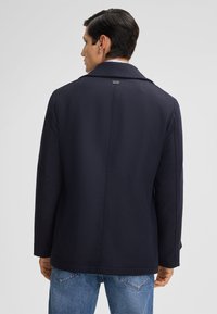 Mann in einem dunkelblauen Peacoat mit breitem Kragen, von hinten gesehen, kombiniert mit hellblauen Jeans vor einem neutralen Hintergrund.