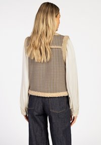 Beige vest met een houndstooth patroon, afgewerkt met zachte fleece, en een rechte snit. Draagt over een crème lange mouw shirt en donkere denim.