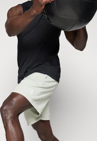 Camiseta sin mangas negra, pantalones cortos verde claro con un logo blanco, brazo musculoso levantando una bola de peso negra, enfoque en los detalles de ropa deportiva.