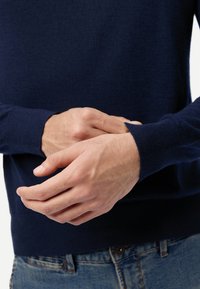 Marineblauer Pullover aus weichem Material, mit einem gerippten Bündchen und einem Rundhalsausschnitt. Die Hände sind nahe am Saum positioniert, wobei das Handgelenkdetail sichtbar ist.