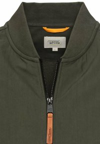 Olijfgroene jas met een staande kraag, voorkant met ritssluiting en contrasterende oranje binnenkant. Voorzien van een beige ritshendel en merklabel.