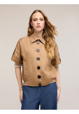 Młoda kobieta o długich, falowanych włosach, ubrana w beżową, krótkorękawową, zapinaną na guziki kurtkę i niebieskie spodnie, stojąca na jednolitym tle.