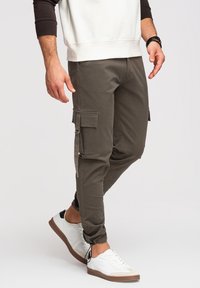 Homme portant un pantalon cargo gris foncé avec des poches latérales et des baskets blanches, marchant sur un fond clair uni.
