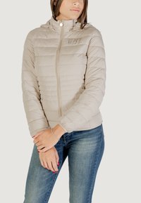 Giacca puffer beige chiaro con cappuccio, design trapuntato, cerniera frontale e logo EA7. Abbinata a jeans blu, che mostrano una texture e una vestibilità lisce.