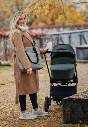Gray quilted tote bag with black straps held in one hand, while the other hand holds a cup near a dark green stroller on a path.