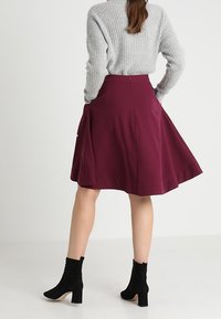 Burgunderfarbener, ausgestellter Rock aus gewebtem Stoff mit glatter Textur, kombiniert mit schwarzen Knöchelstiefeln und einem grau gerippten Strickpullover.