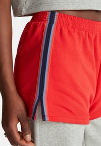 Osoba nosící červené sportovní šortky s modrými a černými svislými pruhy přes šedé tepláky, ruka volně podél těla.