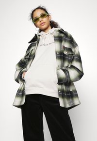 Per didelė žalia plaid flanelinė marškinė, dėvimi virš šviesiai pilkos mikimauzės su užrašu "Abrand Jeans". Juodi kordūro marškiniai ir žali akiniai nuo saulės.