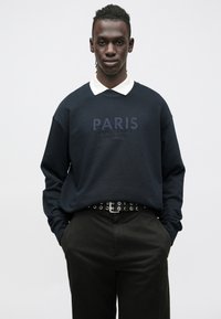 Sudadera azul oscuro con texto "PARIS", combinada con una camisa blanca con cuello. La modelo lleva pantalones negros con un cinturón de ojales. Fondo liso.