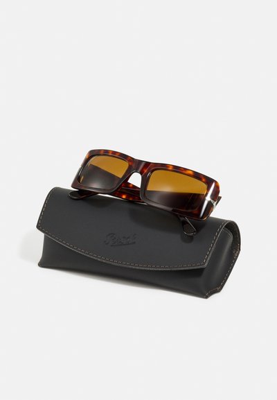 Óculos de sol tartaruga com lentes castanhas a descansar sobre um Estojo de couro preto. O estojo apresenta costura branca e um logótipo.
