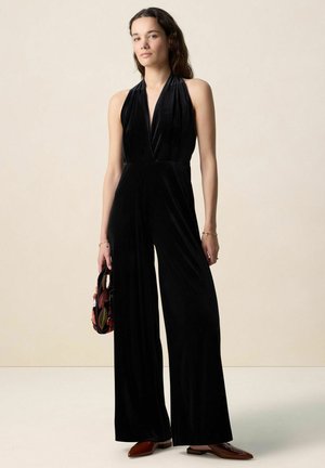 Femme portant une combinaison-pantalon noire en velours sans manches à col noué avec jambes larges, tenant un sac à main coloré en perles et portant des chaussures pointues marron.