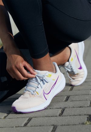 Personne portant un legging noir qui attache les lacets de chaussures de course Nike beige et violettes sur une surface pavée en extérieur.