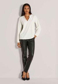 Vrouw in een witte blouse met lange mouwen, zwarte leren broek en zwarte hoge hakken, staande tegen een effen lichte achtergrond.