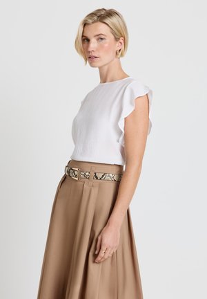 Frau trägt weiße, gerüschte ärmellose Bluse, die in einen hoch taillierten beigen Rock mit Schlangenledergürtel gesteckt ist, steht vor einem schlichten Hintergrund.