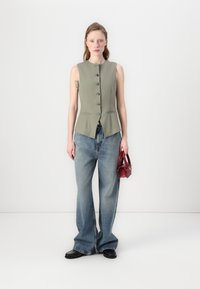 Lys olivengrønn ermeløs vest med knappdetaljer, parret med vide blå jeans og svarte sko, holding en liten rød håndveske.