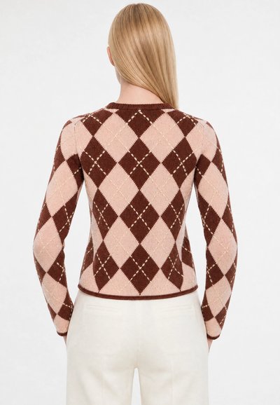 Personne aux longs cheveux blonds vue de dos portant un pull à motif losanges marron et rose et un pantalon blanc.