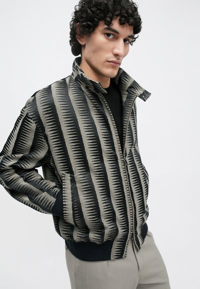 Jacke mit Reißverschluss in Schwarz und Beige mit Zickzackmuster und gerippten Bündchen. Verfügt über Seitentaschen und einen hohen Kragen. Glatte Textur.
