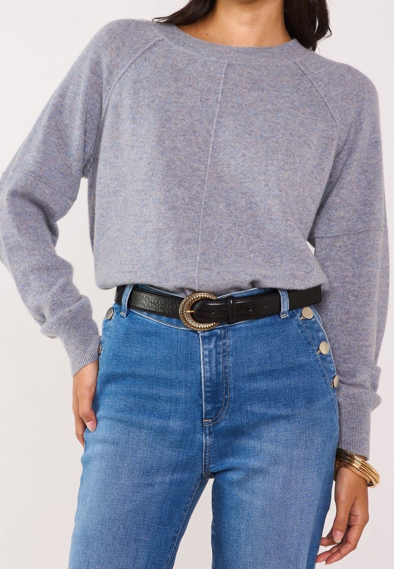 Femme portant un pull en maille gris clair rentrée dans un jean taille haute bleu avec des boutons argentés et une ceinture noire à boucle dorée.