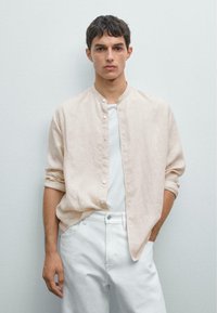 Hellbeige, gestreiftes Hemd mit Knopfleiste und Stehkragen, kombiniert mit einem weißen T-Shirt und weißen Hosen. Glatter Stoff, lockere Passform.
