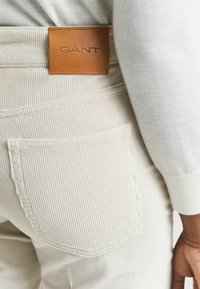 Pantalon en velours côtelé blanc avec une surface texturée, arborant une étiquette en cuir marron embossée avec "GANT" sur la ceinture et une poche arrière.