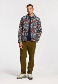 Jachetă cu model floral din material multicolor, bază închisă la culoare, asortată cu pantaloni din catifea de culoare măslinie, adidași gri și cămașă bleumarin pe dedesubt.