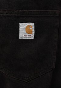 Τσέ πη με λογότυπο Carhartt σε σκούρο ύφασμα, με λευκή ετικέτα που φέρει πορτοκαλί λογότυπο και καφέ κείμενο. Ραμμένες άκρες, υφή με ανάγλυφη επιφάνεια.