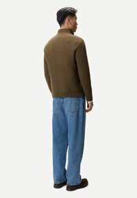 Mann steht mit dem Rücken zur Kamera, trägt einen braunen Rollkragenpullover, lockere blaue Jeans und dunkelbraune Schuhe vor einem weißen Hintergrund.