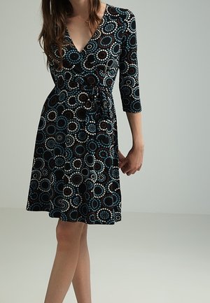 Donna che indossa un vestito nero al ginocchio con motivi circolari blu, bianchi e marroni e maniche a tre quarti, in piedi davanti a uno sfondo grigio uniforme.