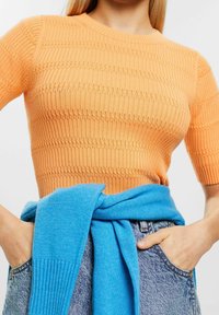 Oranžový žebrovaný pletený top s krátkým rukávem a kulatým výstřihem, doplněný modrým svetrem uvázaným v pase a džíny.