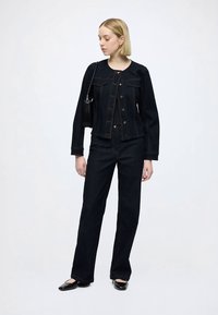 Mörk denimjacka med knappstängning, två bröstfickor och matchande mörka denimbyxor. Modellen har en liten svart väska som accessoar.