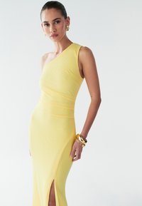 Ein asymmetrisches, gelbes Kleid aus strukturiertem Stoff, mit einem seitlichen Schlitz und figurbetontem Schnitt, ergänzt durch ein goldenes Armband und Ohrringe.