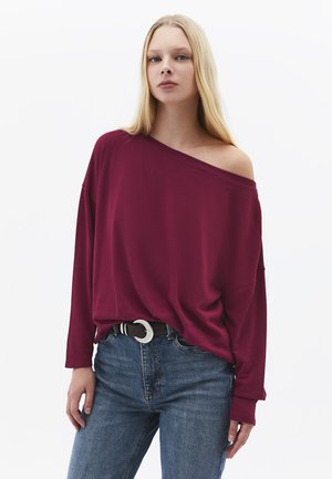 Blonde vrouw die een losse bordeauxrode off-shoulder top draagt, gestopt in een blauwe spijkerbroek met een zwarte riem, staand tegen een witte achtergrond.