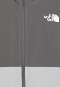 Giacca grigia con la parte inferiore di un grigio più chiaro, dotata di una zip completa e logo bianco in alto a sinistra. Texture liscia, senza motivi.