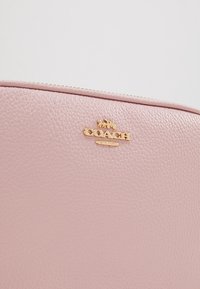 Vaaleanpunainen nahkabagi, jossa on teksturoitu pinta, kultaiset yksityiskohdat ja kohokuvioitu "Coach" -logo. Vetoketjullinen yläosa.