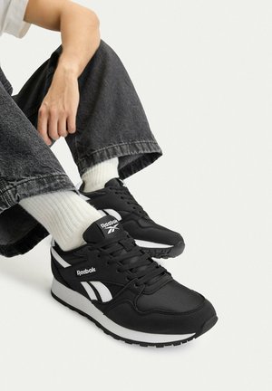 Osoba nosząca czarne sneakersy Reebok, białe skarpetki i ciemne, szerokie spodnie, siedząca z nogami zgiętymi na jednolitym tle.