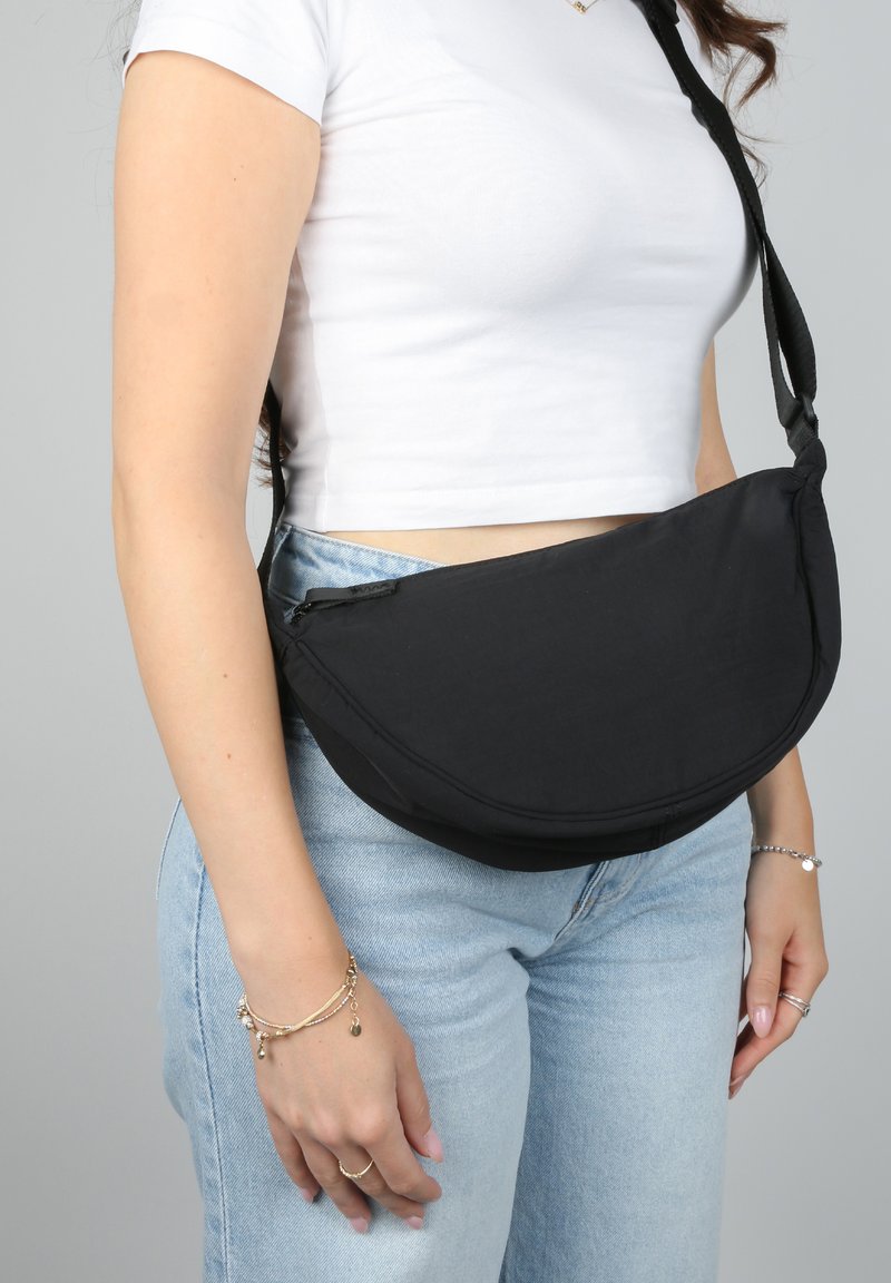 Donna in un crop top bianco e jeans blu chiaro con una borsa a tracolla nera appoggiata sul fianco, su uno sfondo grigio semplice.