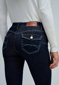 Vaqueros de mezclilla oscura con un diseño ajustado. Presenta una etiqueta de cuero marrón, detalles en hardware dorado y costuras blancas contrastantes en los bolsillos.