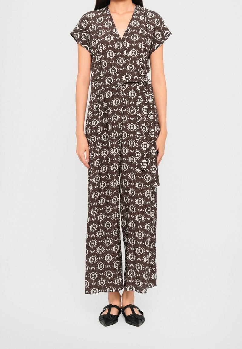 Femme portant une combinaison brun à motifs avec des manches courtes, des jambes larges, un col en V et des ballerines pointues noires.